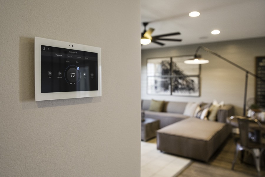 A climate control touchscreen interface mounted on a wall with a living room in the background.